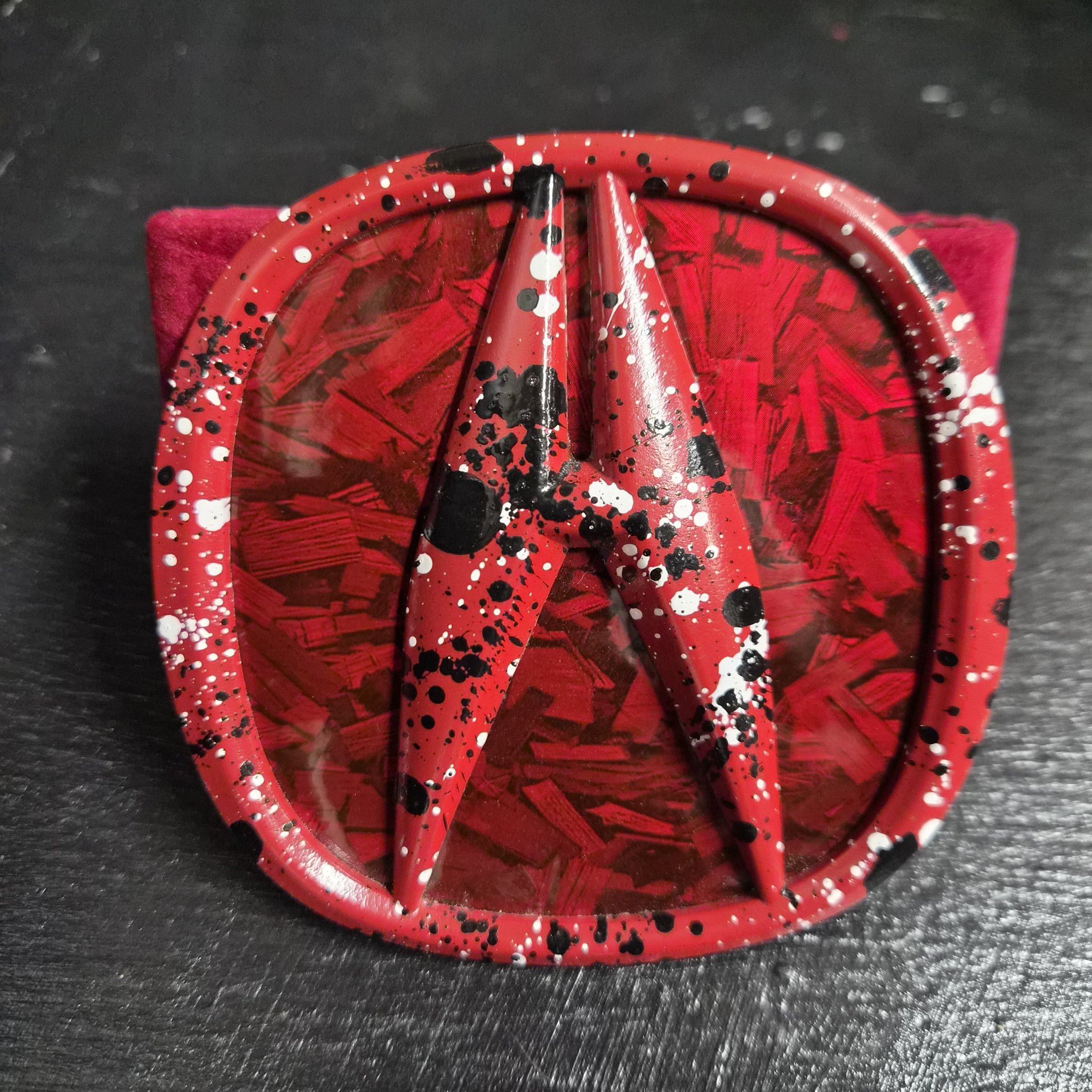 Front-on close-up of a red-splatter Acura emblem on a forged-carbon insert